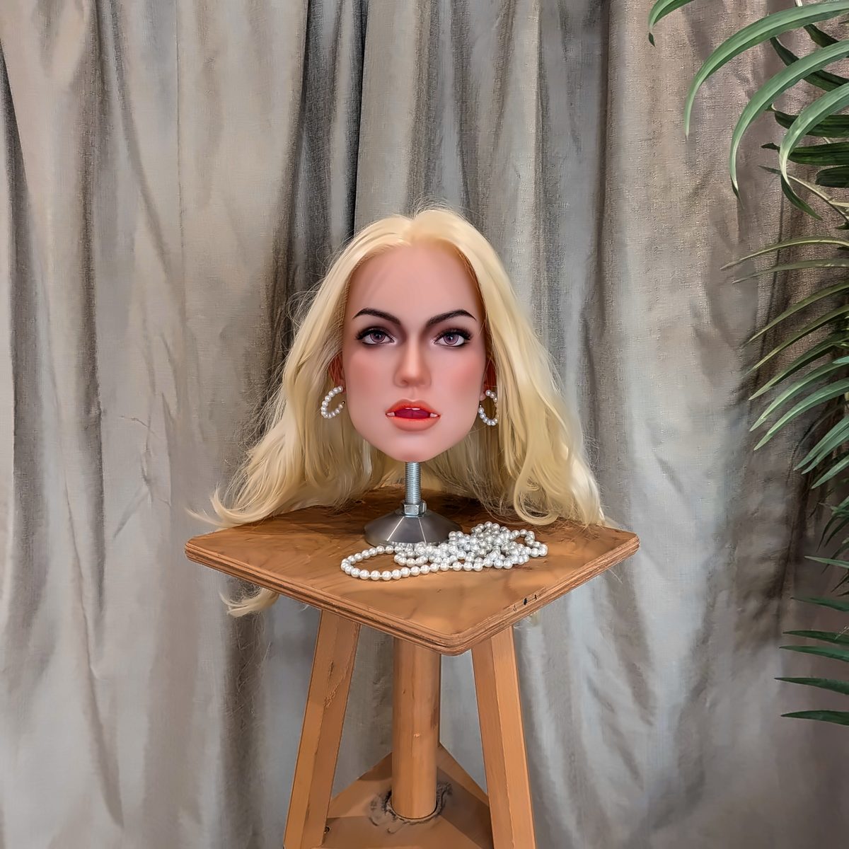 The Blonde look on a wooden stool in front of a grey linen curtain, with a palm leaf at the edge of the frame.