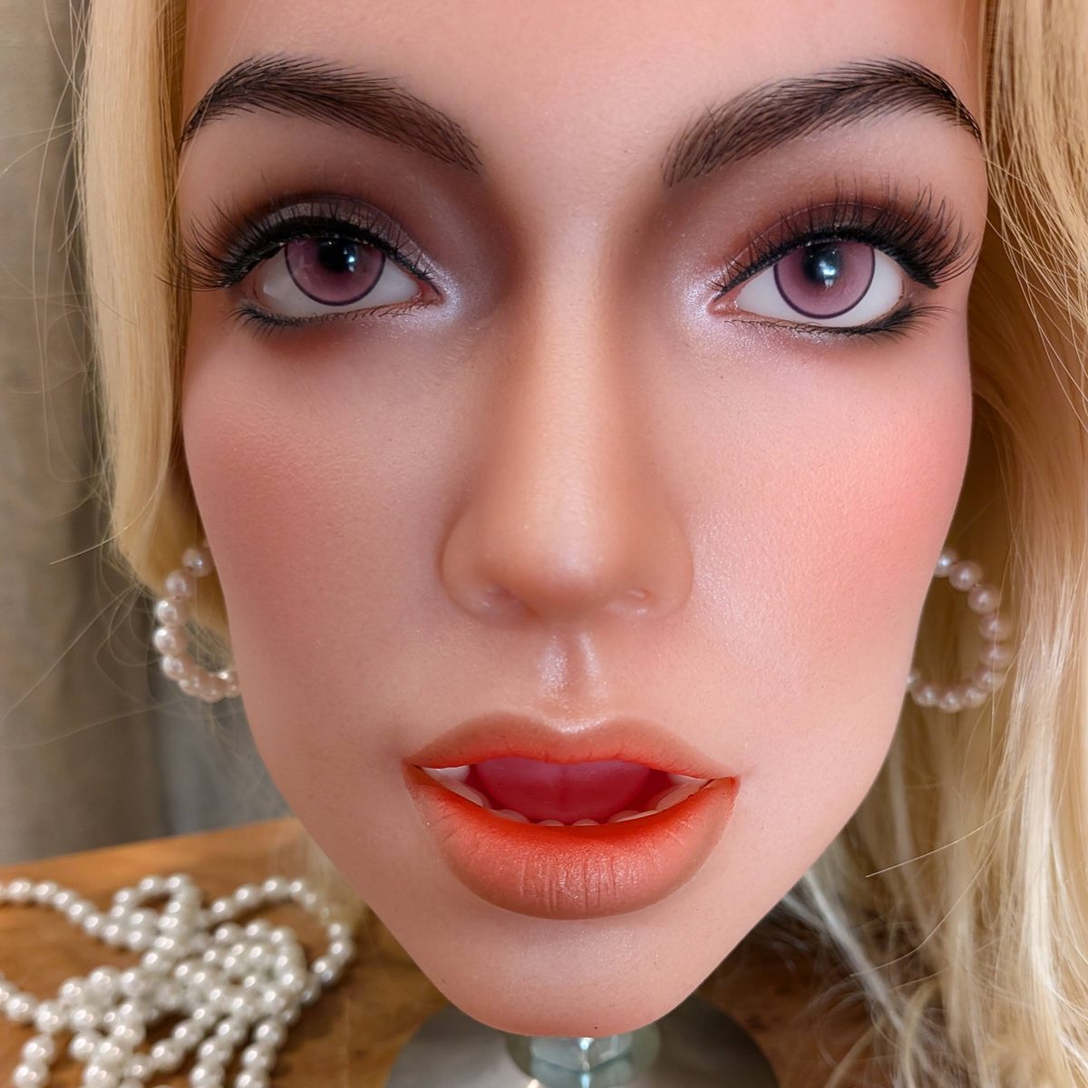Close-up of a face with violet eyes, defined brows, parted coral lips and pearl earrings, with a string of pearls below.