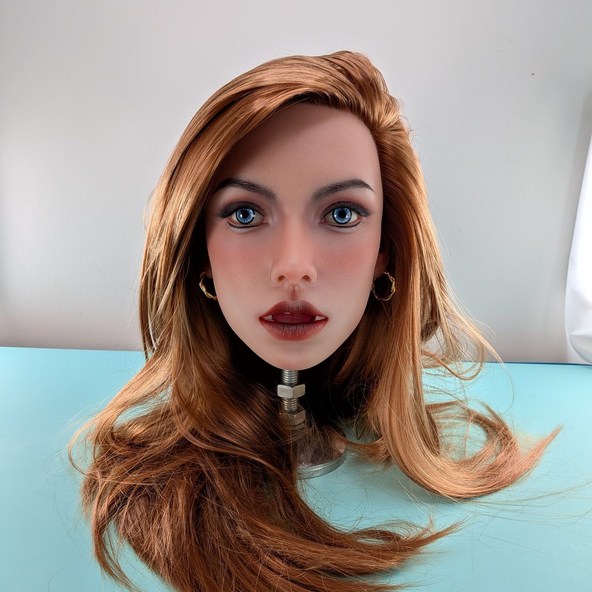 A lifelike head with long copper hair, blue eyes and small gold hoop earrings, on a stand on a turquoise studio table against a white backdrop.