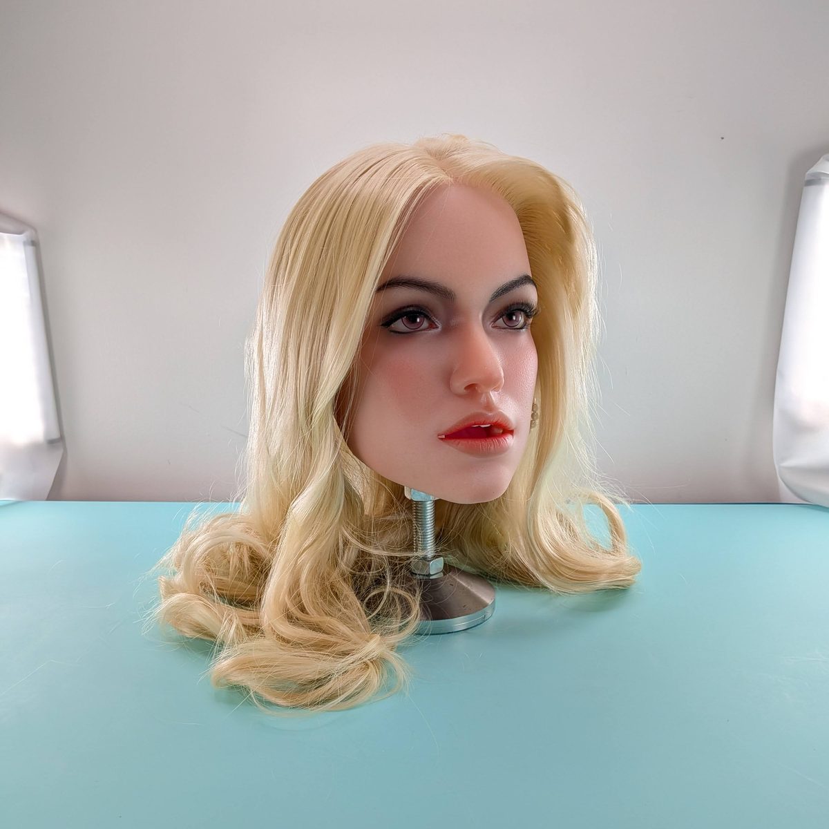 A lifelike head with loose, wavy blonde hair and red lips on a stand on the turquoise studio table.