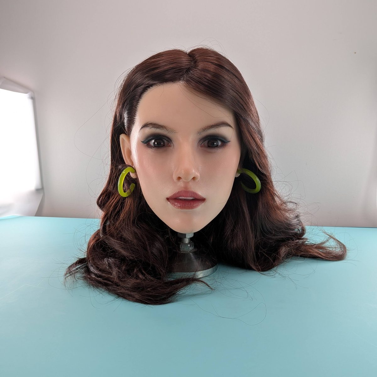 A lifelike head with dark wavy hair and green hoop earrings on the turquoise studio table.