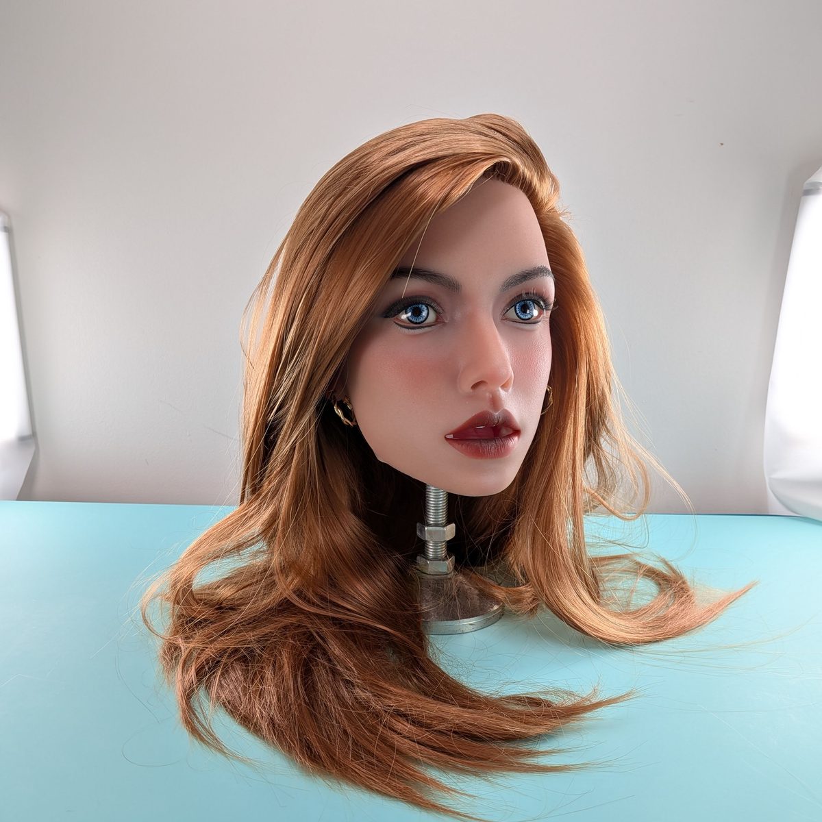 A lifelike head with long, straight auburn hair and blue eyes on the turquoise studio table.