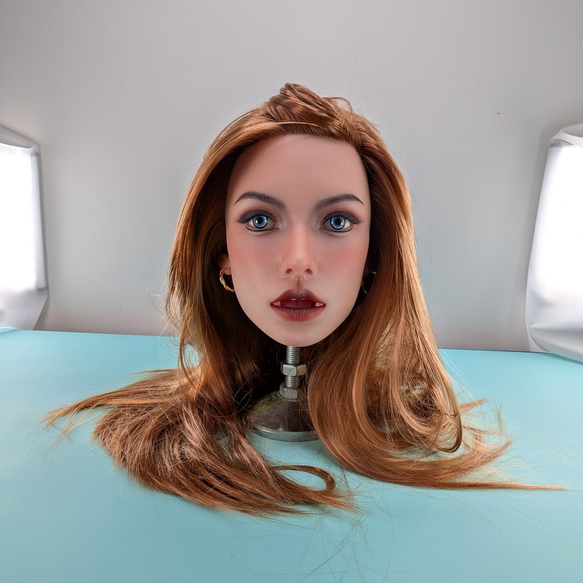 A lifelike head with warm ginger hair, seen in three-quarter profile on the turquoise studio table.